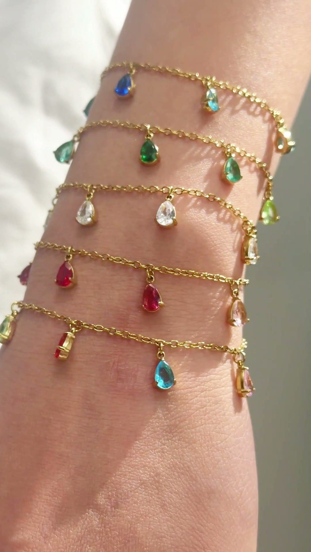 Delicate gold-tone chain bracelets with colorful teardrop-shaped dangling stones shown stacked on the wrist.