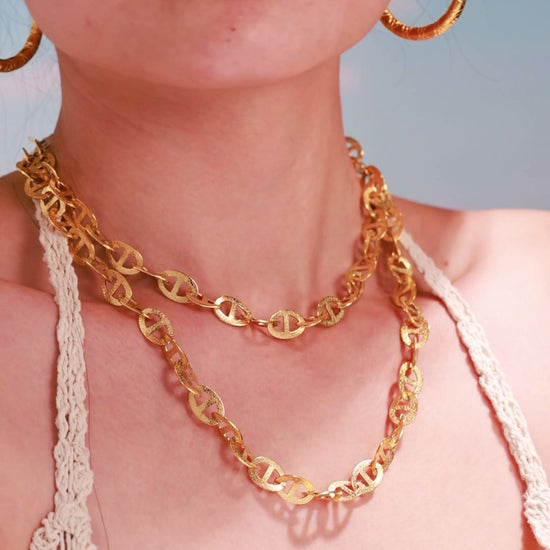 Model wearing layered gold-tone mariner-link necklaces with textured oval links and hoop earrings.