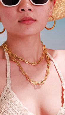 Model wearing layered gold-tone mariner-link necklaces with textured oval links and hoop earrings.