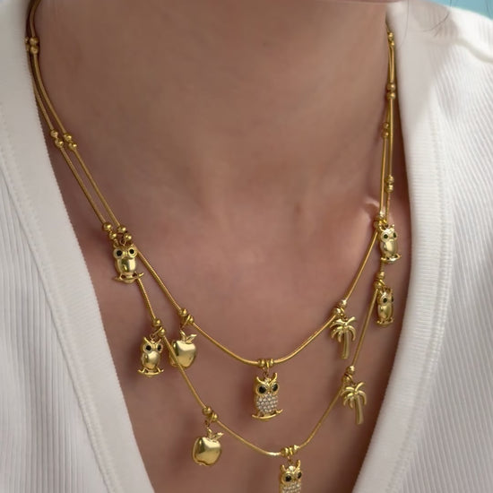 Layered gold-tone charm necklaces with owls, apples and palm trees, featuring clear stone accents, shown on a model.