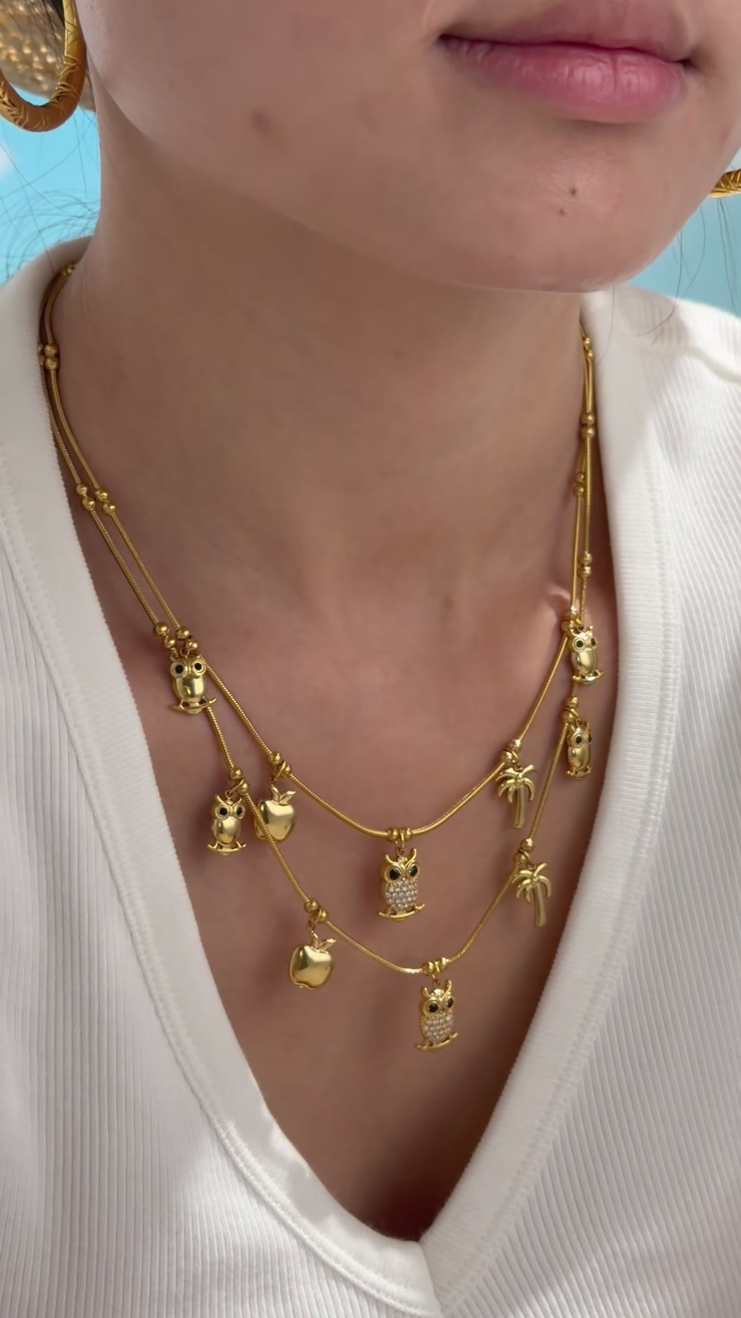 Layered gold-tone charm necklaces with owls, apples and palm trees, featuring clear stone accents, shown on a model.