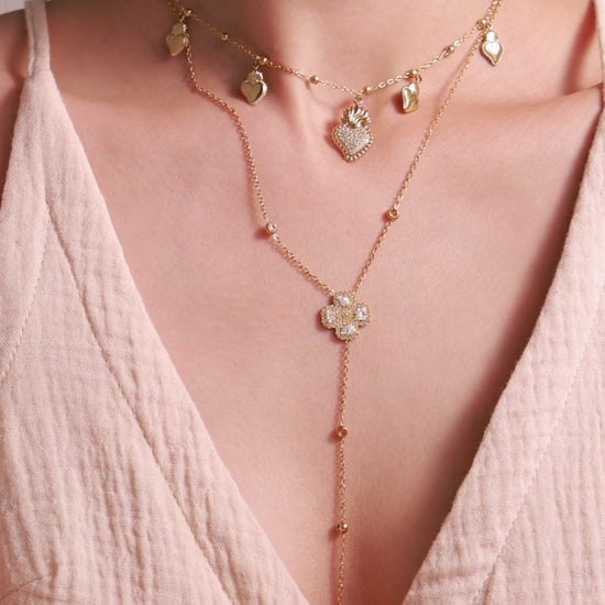 Layered gold-tone necklaces featuring heart charms and a clear-stone clover lariat, with matching bracelets on a model.