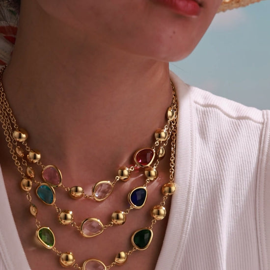 Model wearing layered gold-tone necklaces with colorful faceted stones and polished gold-tone beads.