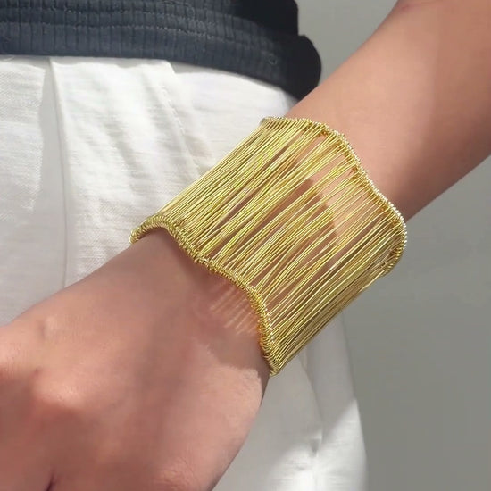 Wide gold-tone openwork cuff bracelet with fine vertical bars and gently curved edges shown on the wrist.