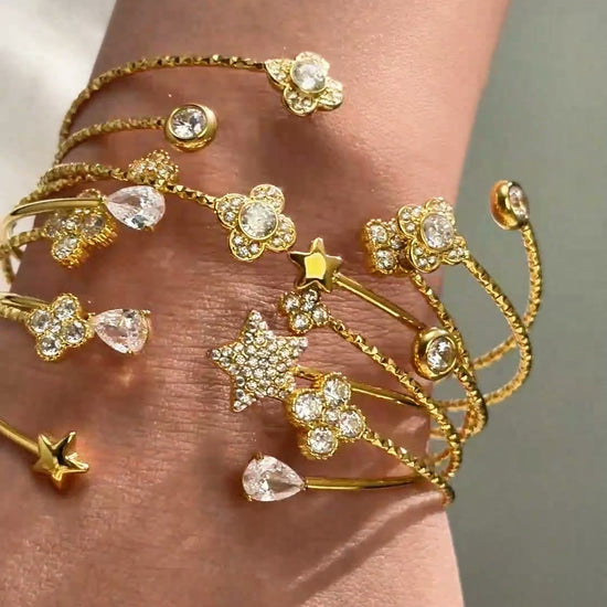 Gold-tone open cuff bracelets with sparkling stars, floral details and clear stones shown stacked on the wrist.