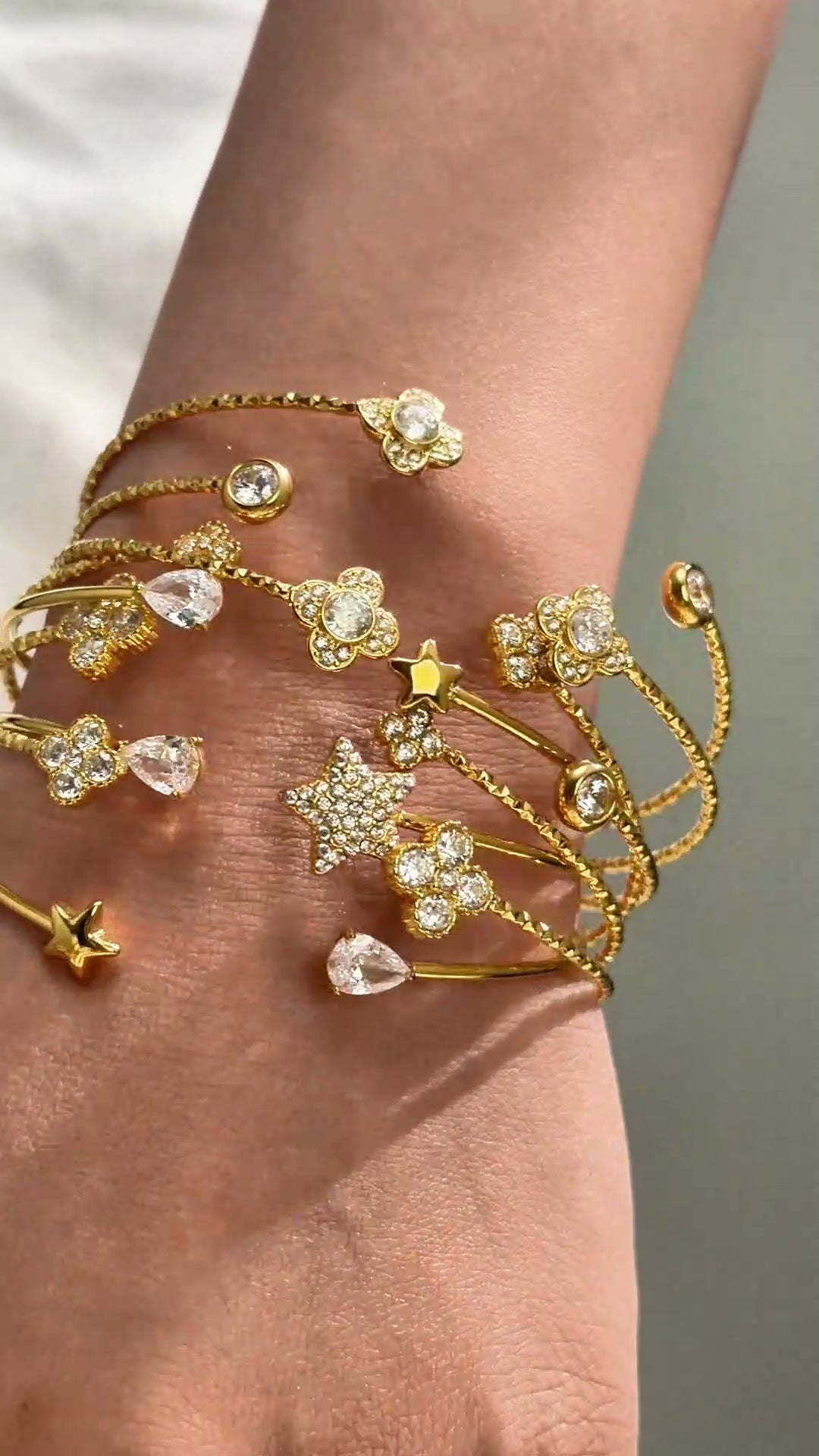 Gold-tone open cuff bracelets with sparkling stars, floral details and clear stones shown stacked on the wrist.