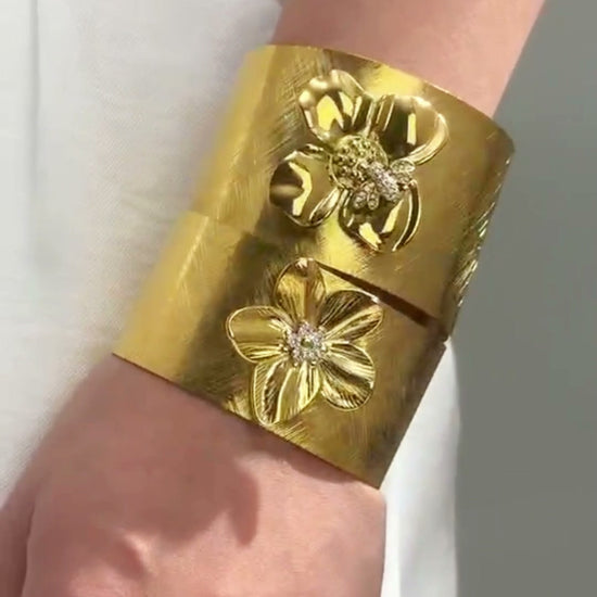 Wide gold-tone cuff bracelets with raised floral details shown stacked on the wrists.
