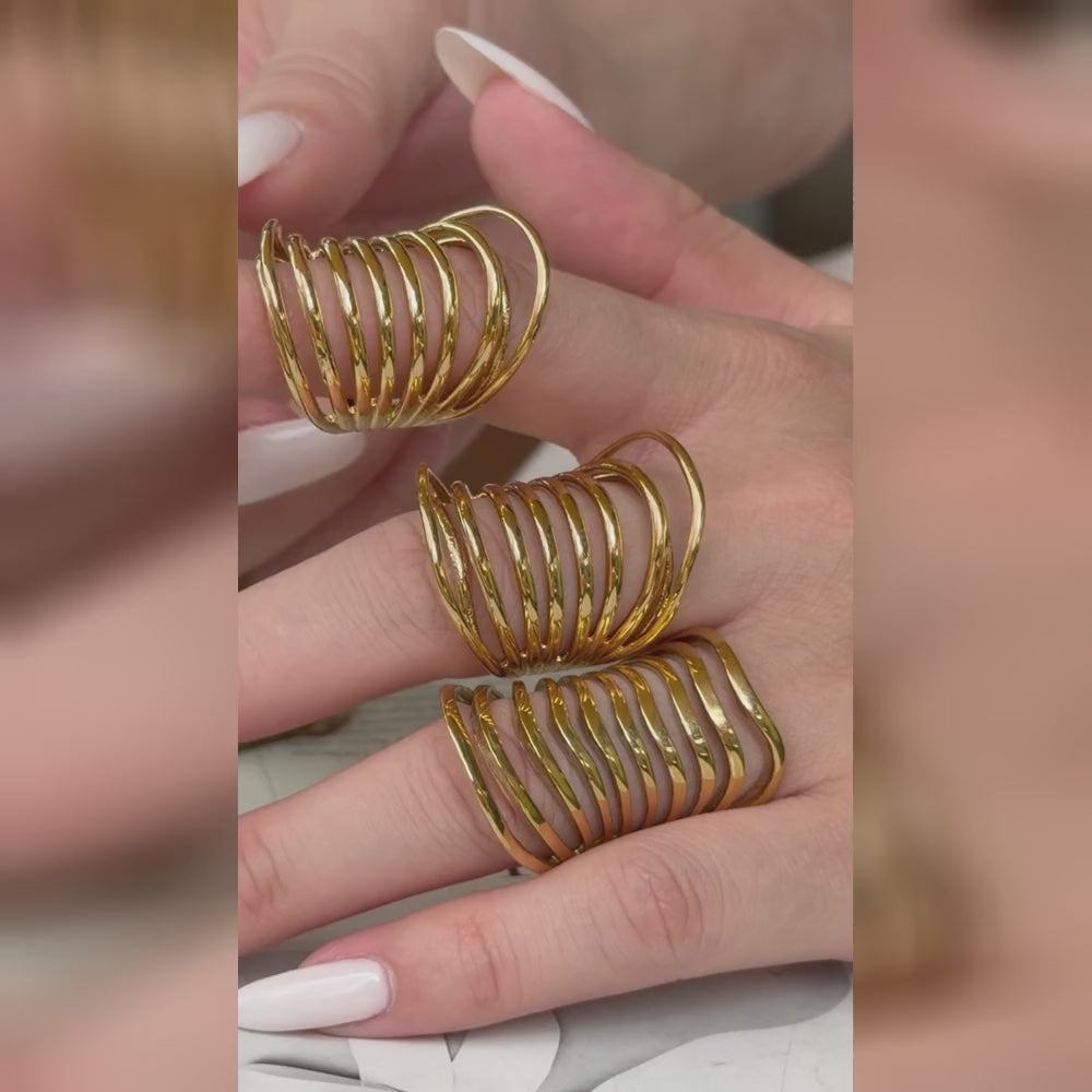 Three wide gold-tone openwork rings with multiple curved bands, shown on a hand with a white manicure.