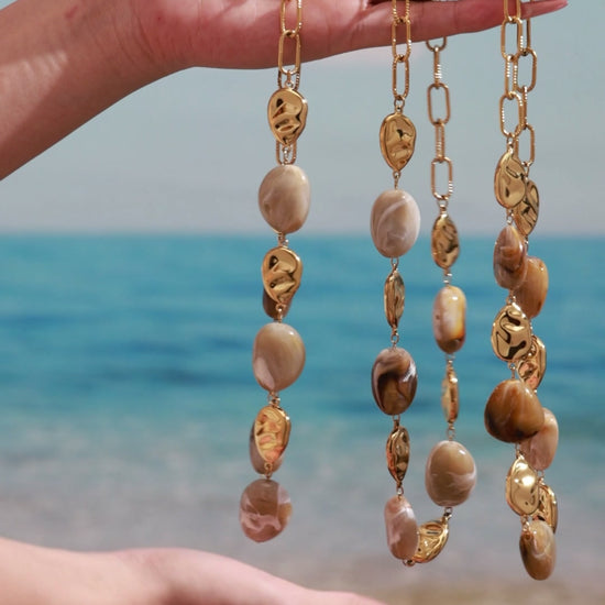 Gold-tone paperclip chain necklaces with brown marbled oval beads and sculpted metal accents, displayed by hand at the beach