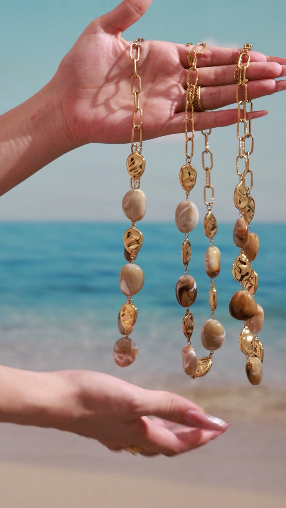 Gold-tone paperclip chain necklaces with brown marbled oval beads and sculpted metal accents, displayed by hand at the beach