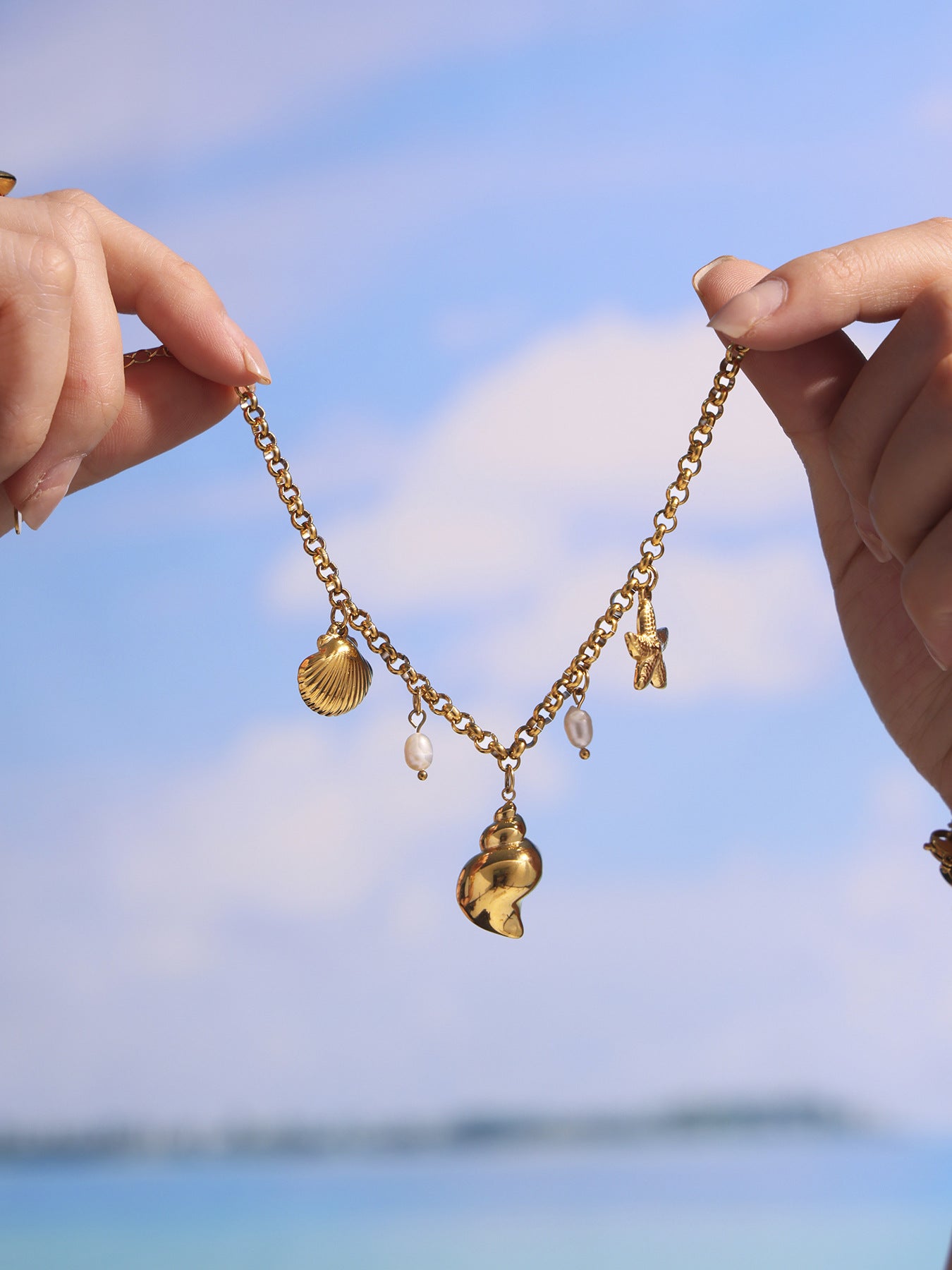 Hand holding gold shell pearl charm anklet against blue beach sky, adjustable stainless steel chain for vacation styling