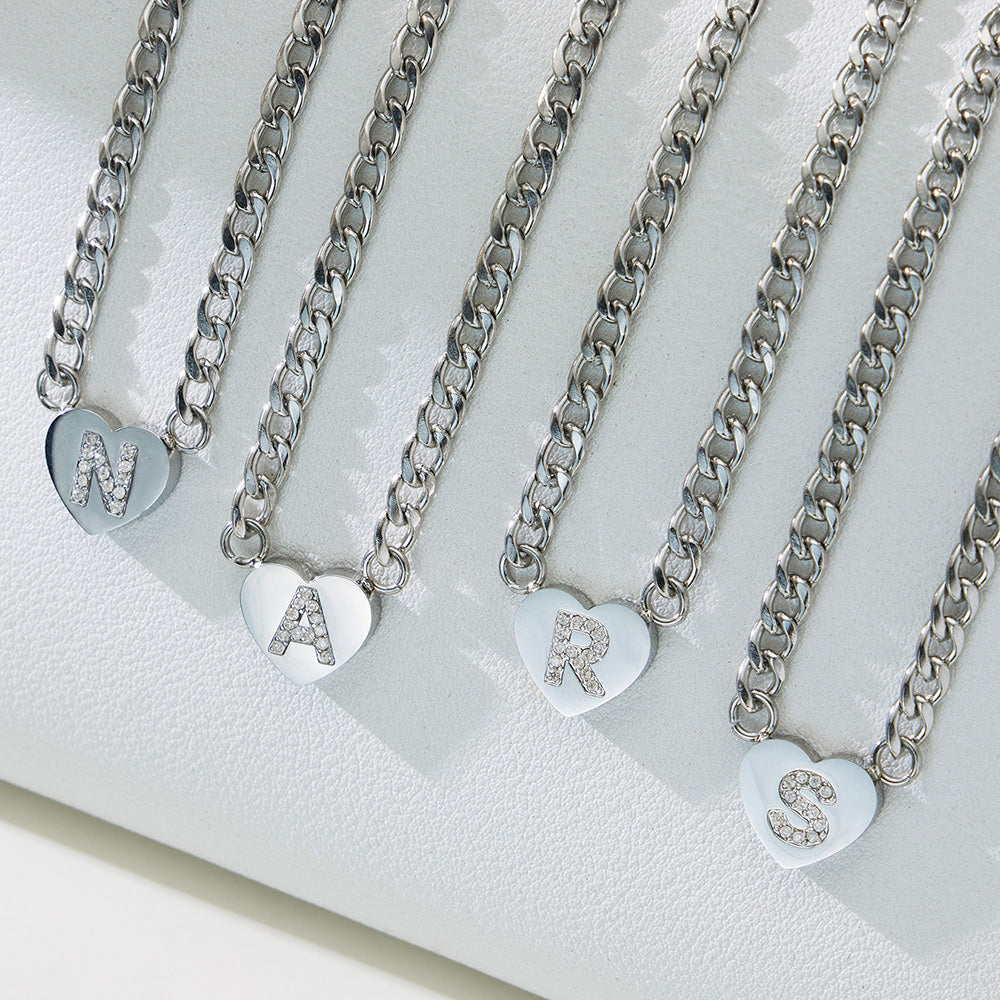 Silver-tone zircon initial heart necklaces displayed in multiple letter variants on a white jewelry stand