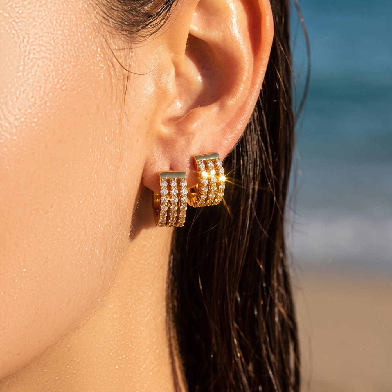 Model wearing compact gold zircon huggie earrings against a beach background
