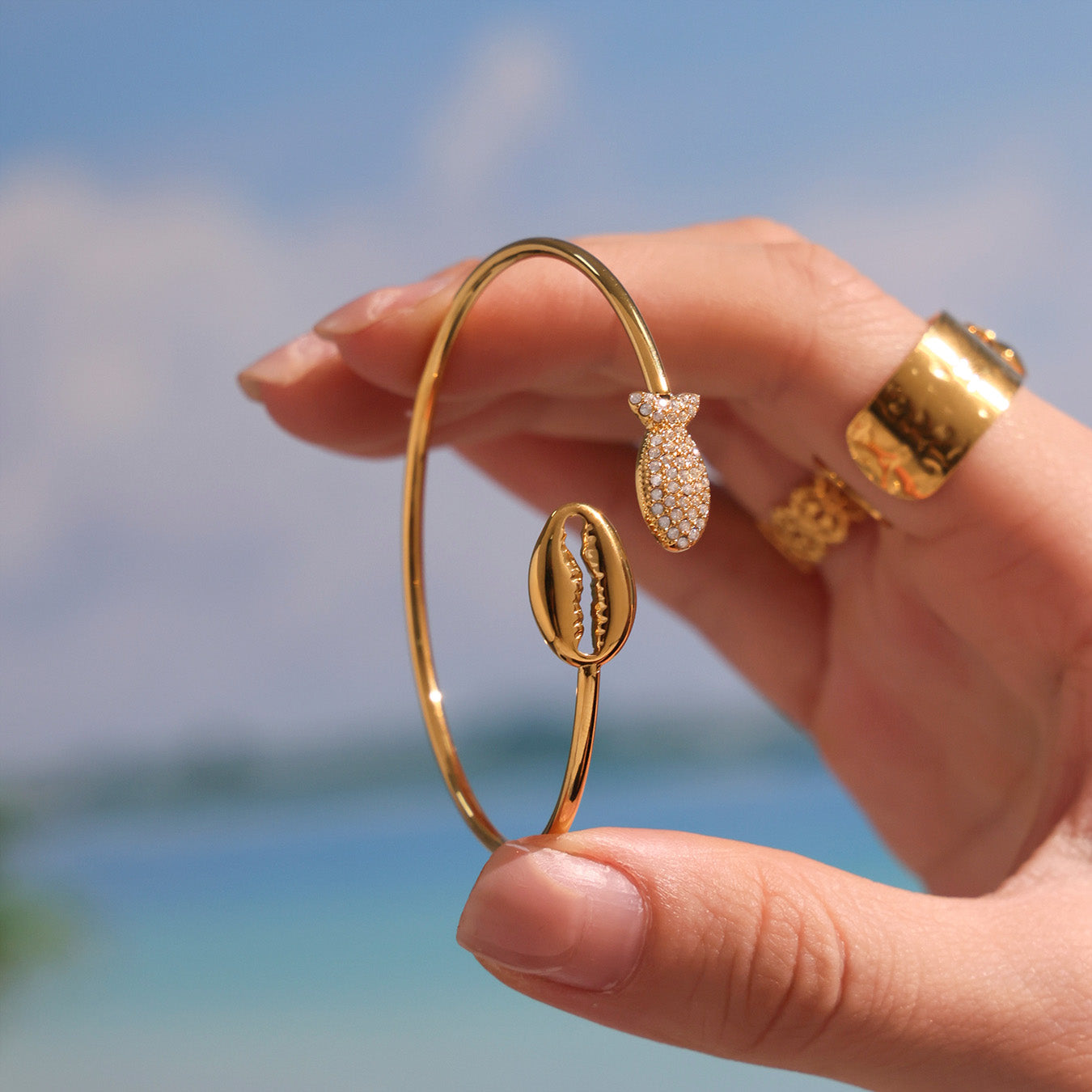 18K gold plated sparkling zircon shell thin open bangle, held in hand against beach background