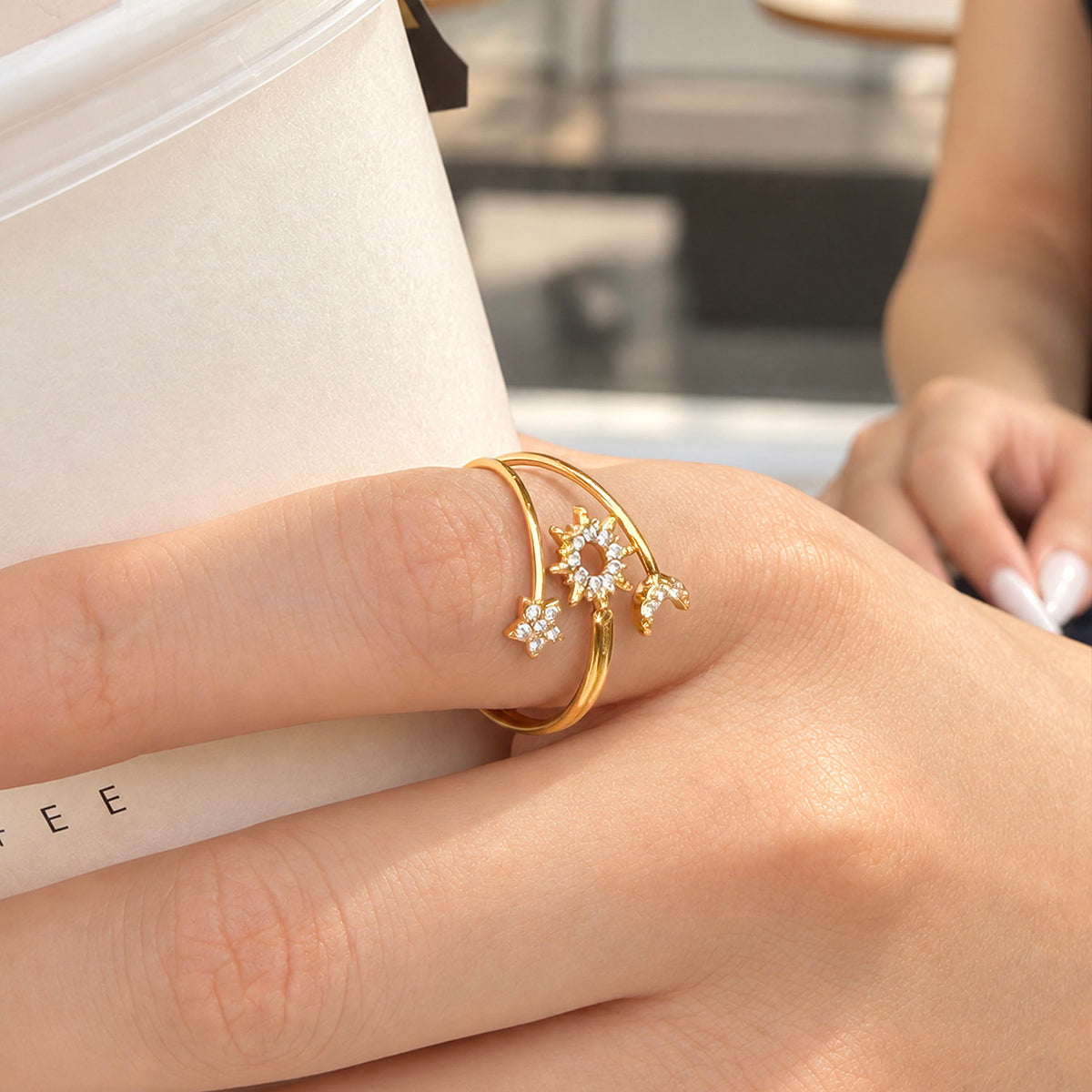 A466 gold-tone stainless steel sun, moon and star open ring worn on a finger beside a white cup