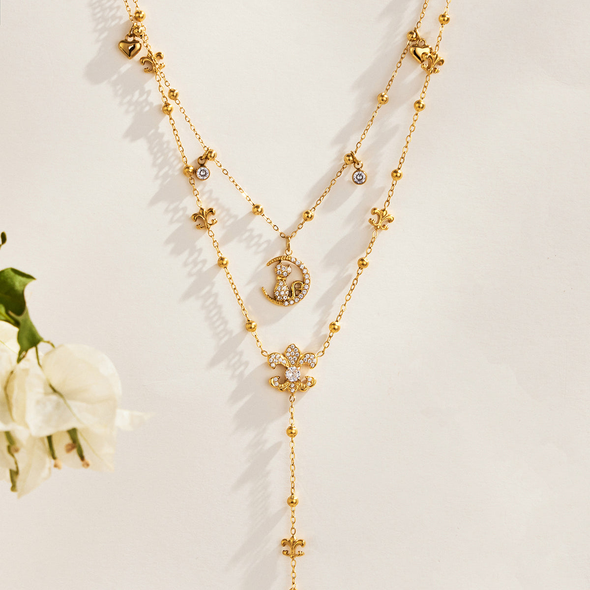Gold-tone stainless steel cat moon pendant and Y lariat necklaces displayed in layers beside white flowers