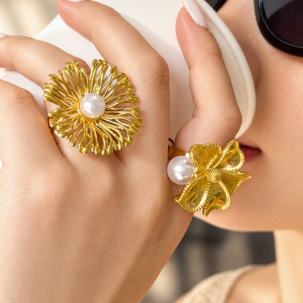 Gold-tone stainless steel openwork and sculpted flower rings with faux pearls worn on fingers held near the face