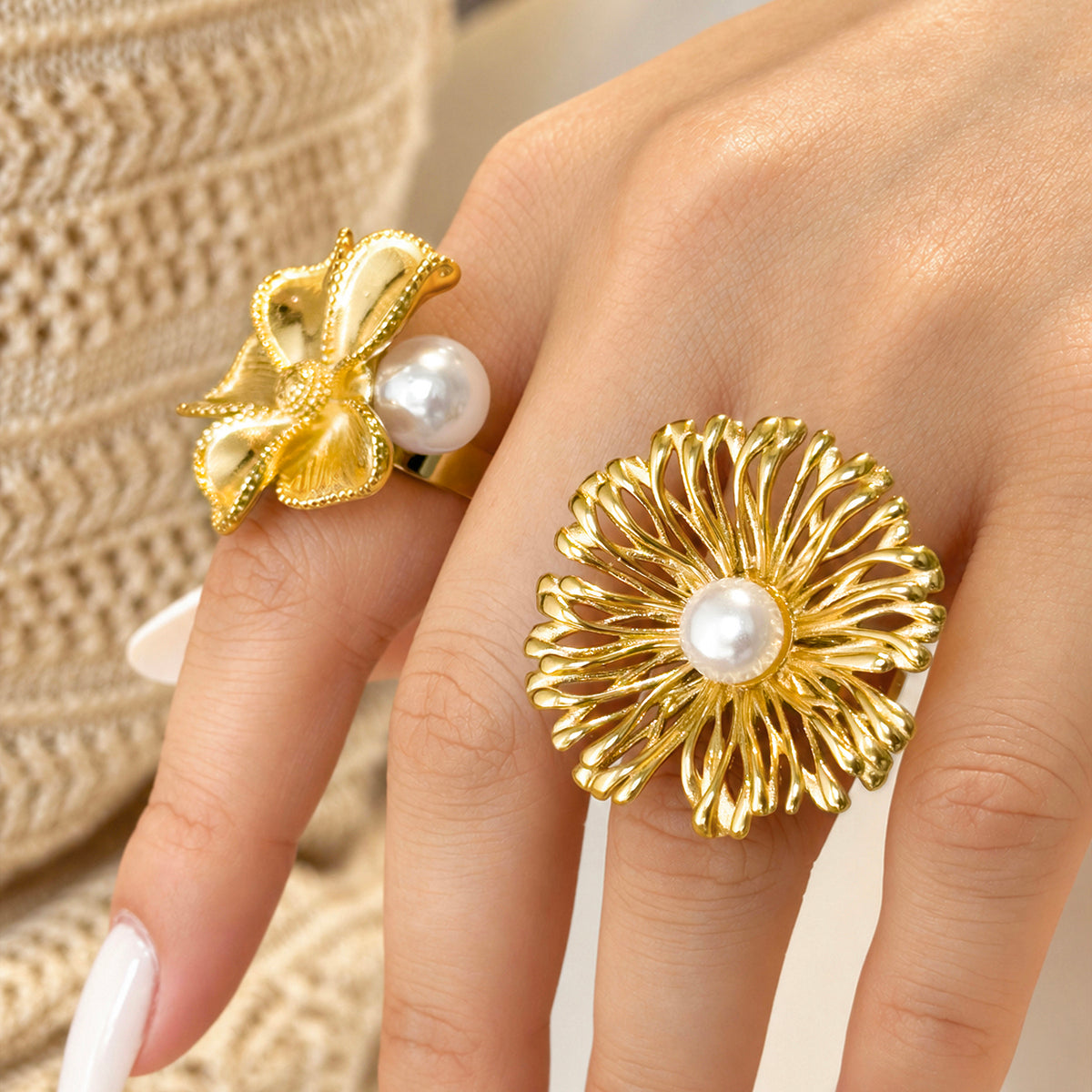 Two gold-tone stainless steel flower rings with white faux pearl details worn against a woven straw background