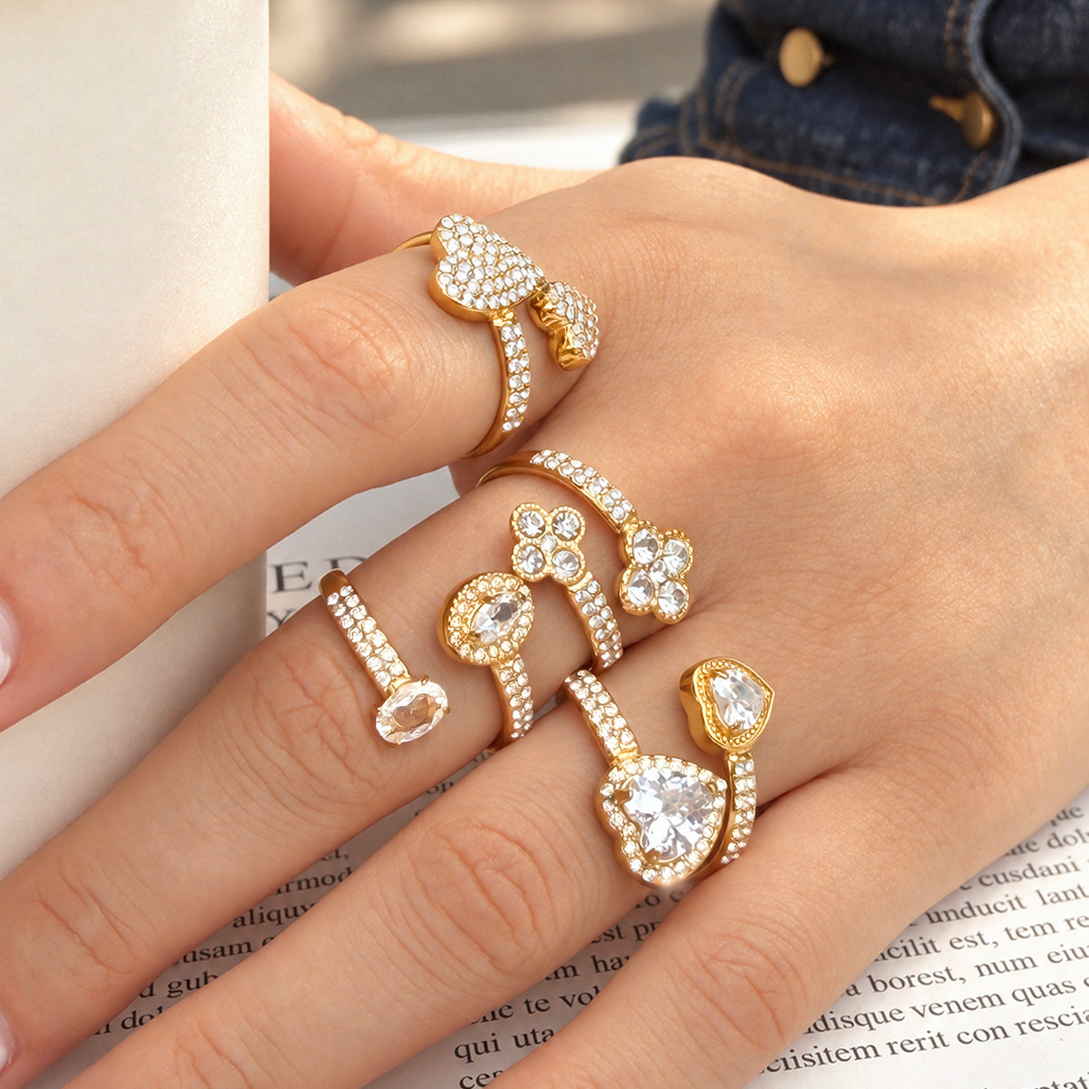 Four gold-tone open rings styled on a hand beside denim and a printed page