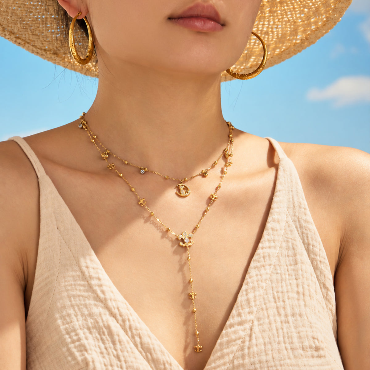 Model wearing layered gold-tone stainless steel cat moon and Y lariat necklaces with a straw hat and cream V-neck top