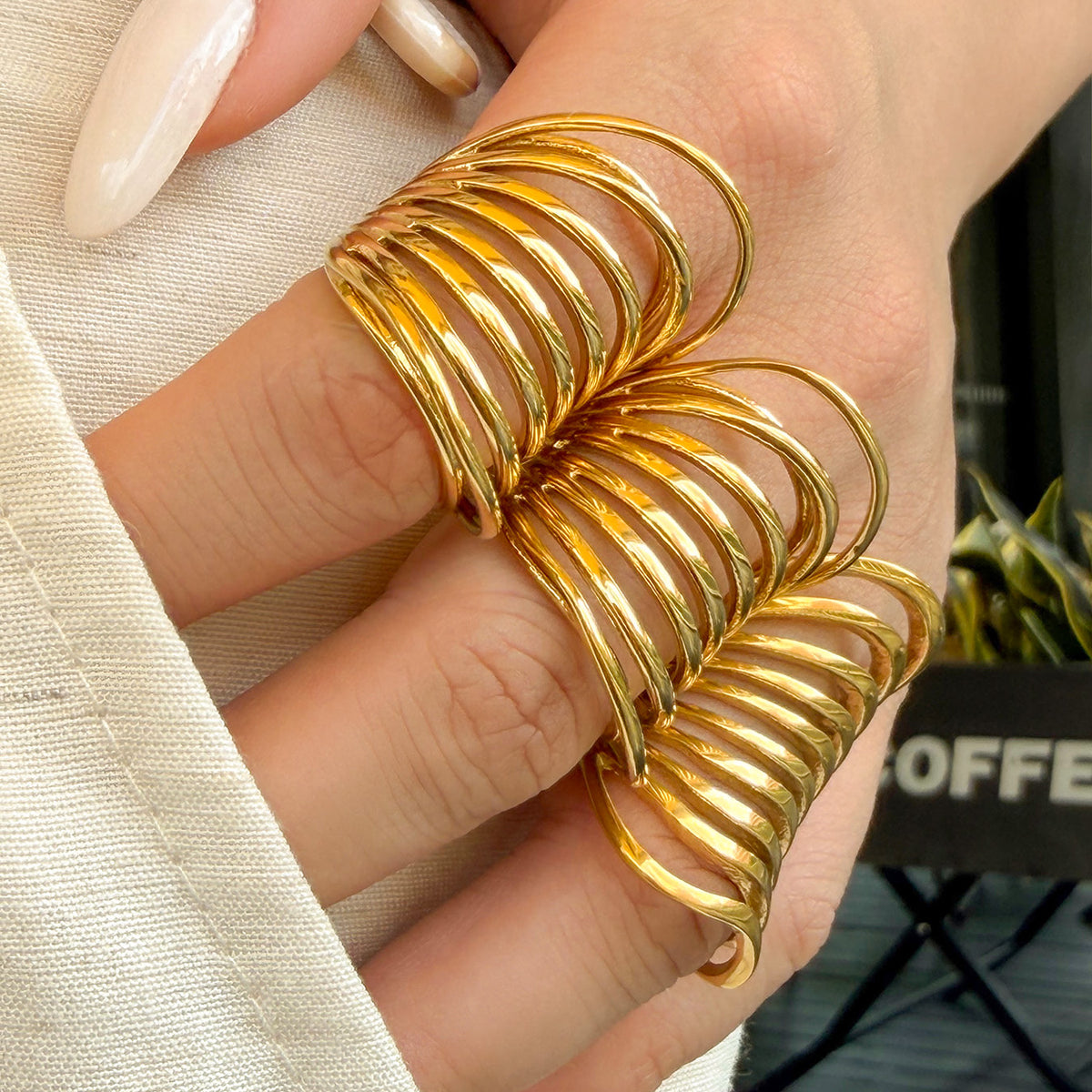 Close-up of gold-tone stainless steel sculptural rings worn on fingers beside a white knit sleeve