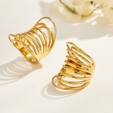Two gold-tone stainless steel sculptural rings on a cream surface, casting shadows through their openwork bands