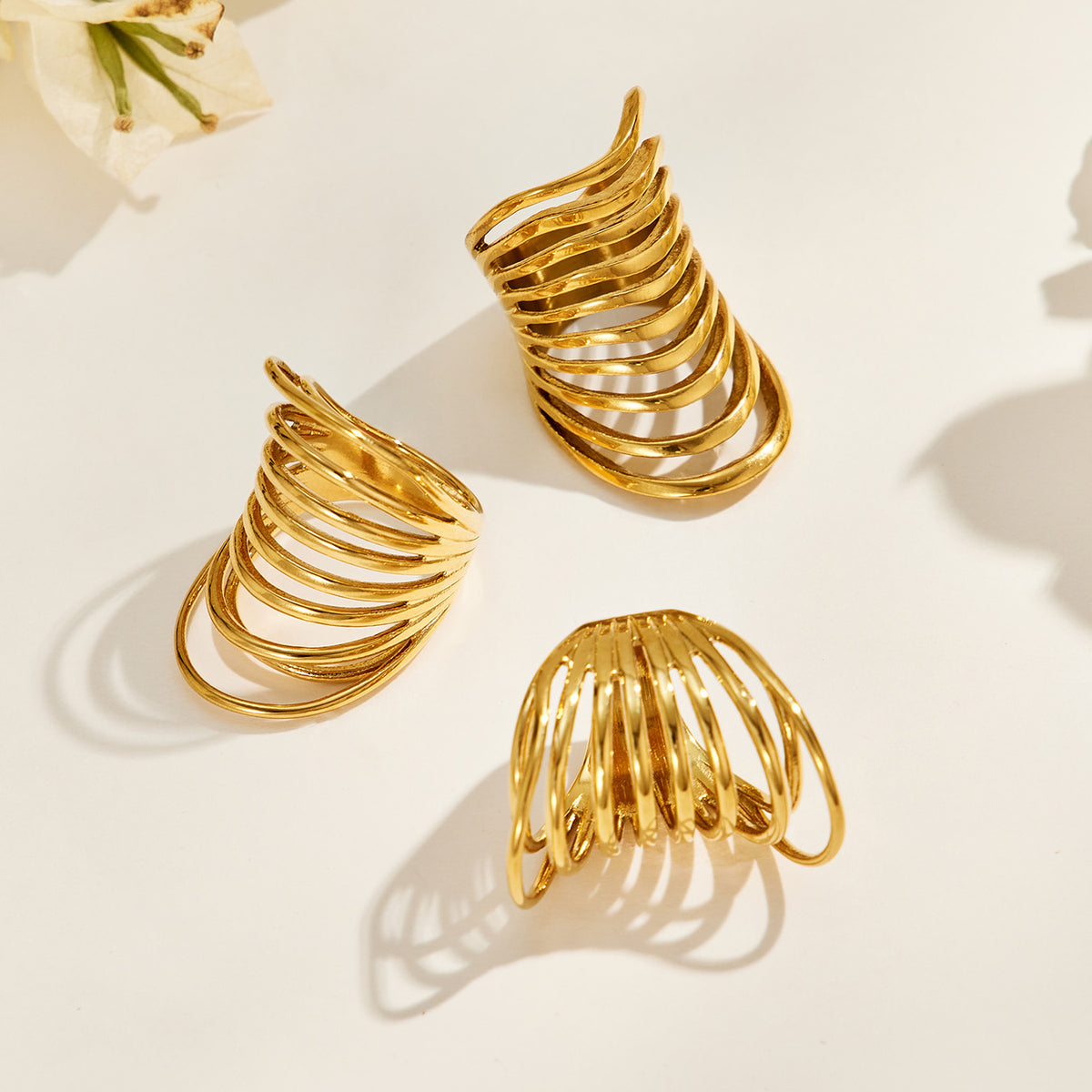 Three gold-tone stainless steel multi-band rings arranged at different angles on a sunlit cream surface