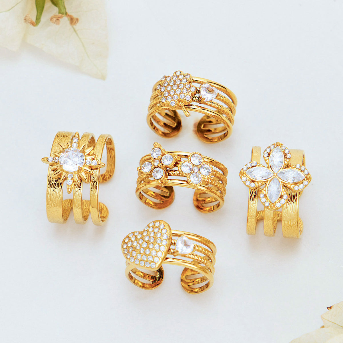 Gold-tone stainless steel statement rings arranged on a cream surface, showing floral, heart and leaf motifs