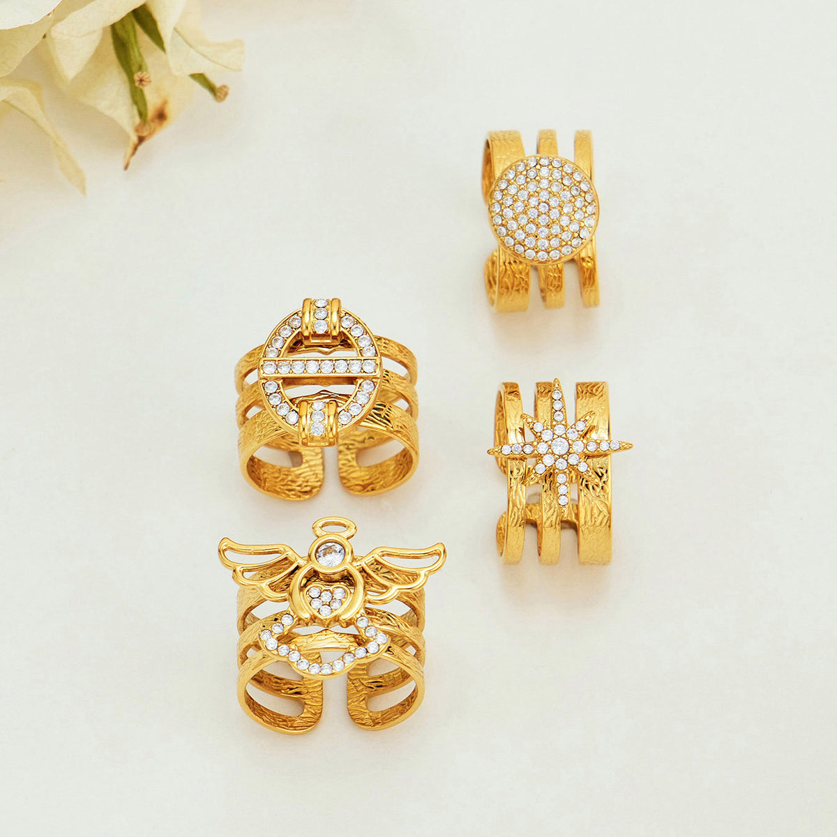Four gold-tone stainless steel rings arranged on a light surface with their geometric, angel, circle and starburst faces visible