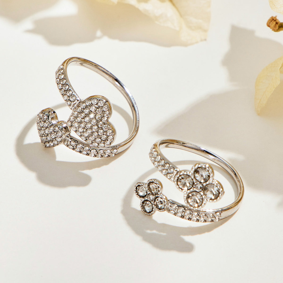 Steel-tone double-flower and pave-heart open rings displayed side by side on a cream surface