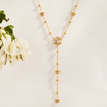 Gold-tone stainless steel Y lariat necklace with ornamental motifs and a beaded chain drop beside white flowers
