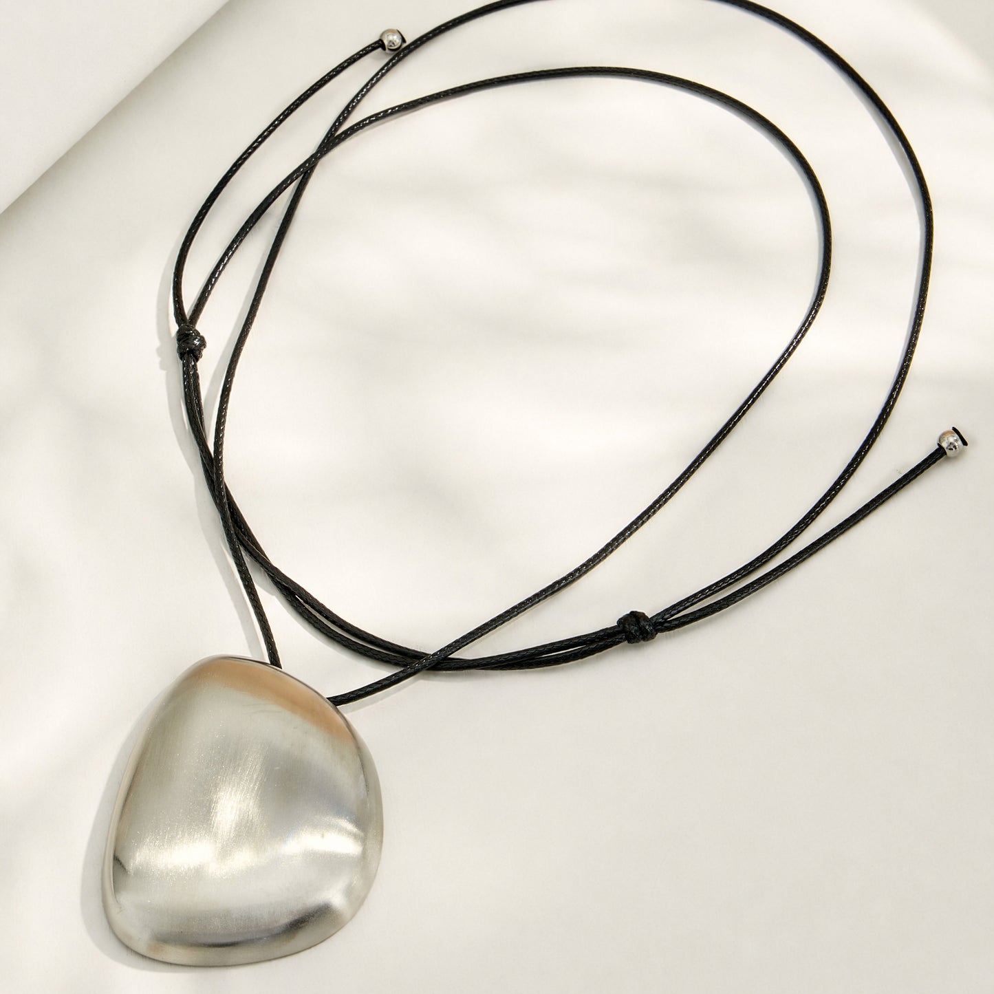 Polished steel pebble pendant necklace with a long black cord arranged in a flat lay