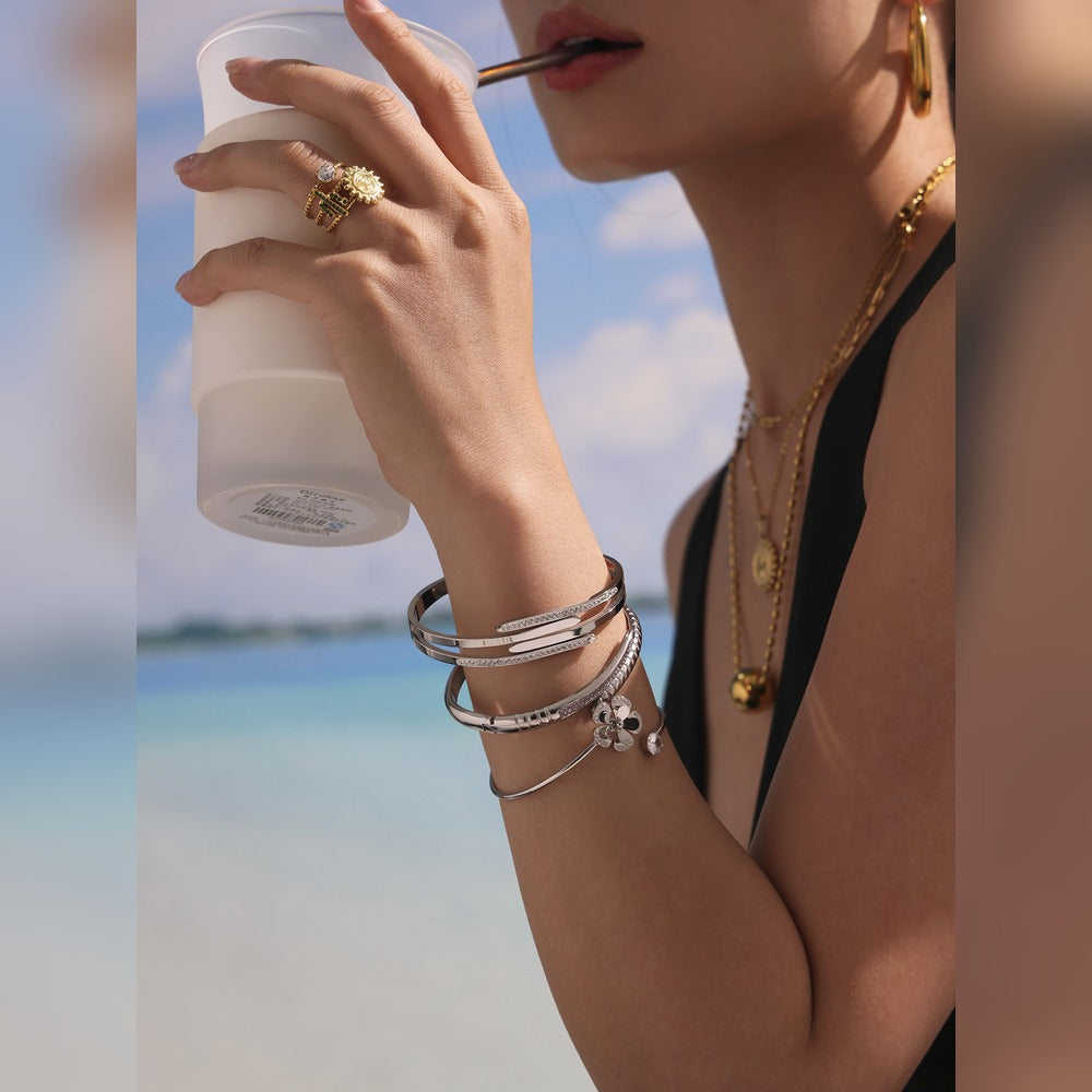 Model wearing a steel-tone nail bangle bracelet stack while holding a drink by the sea