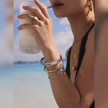 Model wearing a steel-tone nail bangle bracelet stack while holding a drink by the sea