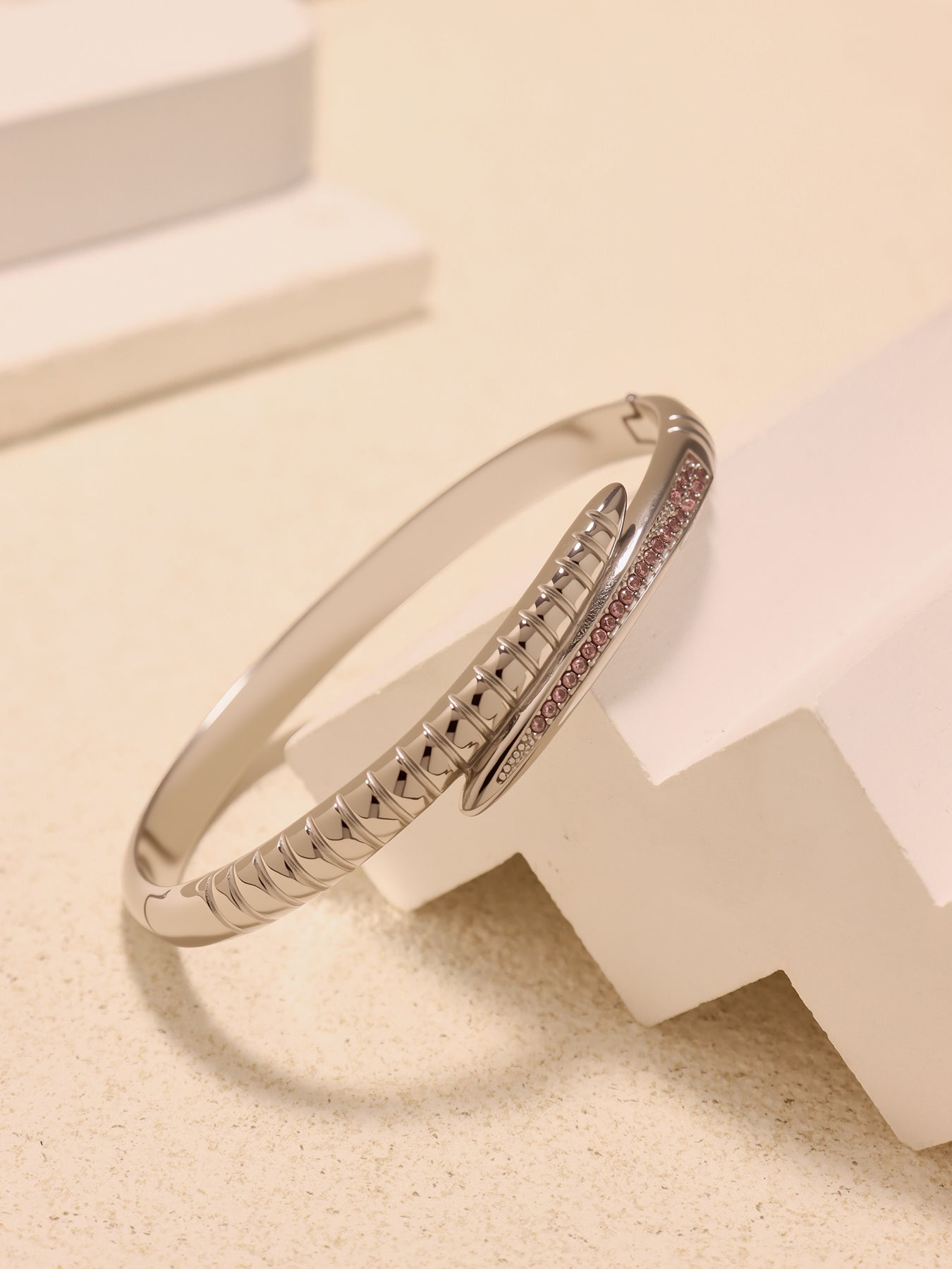 Steel-tone nail bangle bracelet with pink zircon accents displayed on white geometric blocks