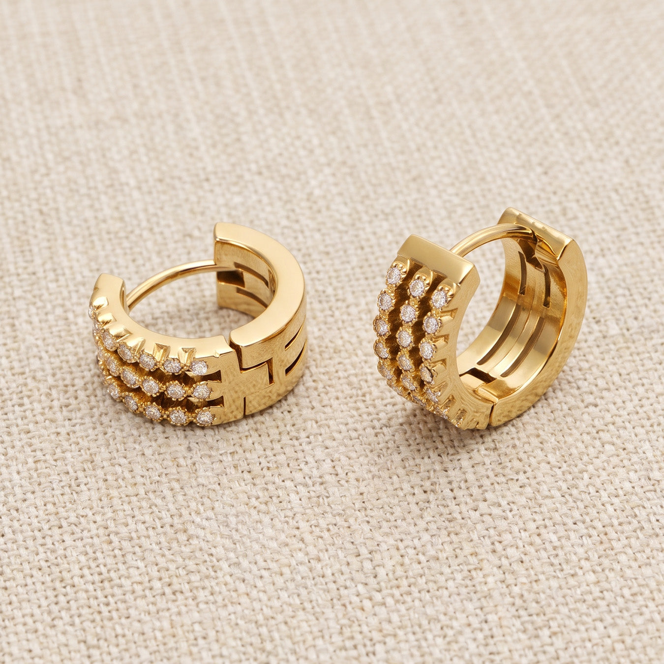 Pair of gold triple-row pavé huggie earrings displayed on woven beige fabric