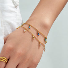 Gold-tone chain bracelets with colorful dangling stone charms styled on a hand with gold rings.