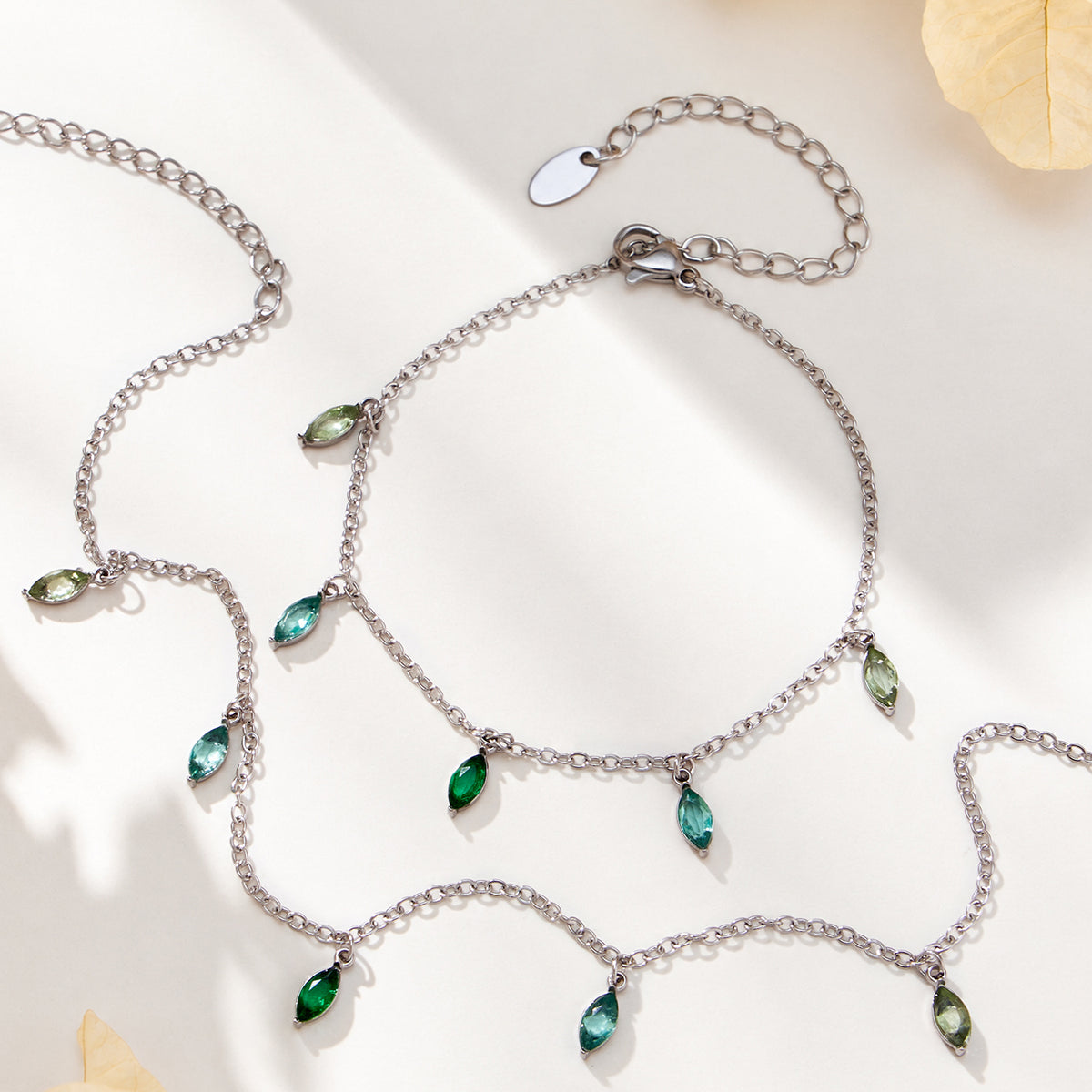 Steel-tone fine-chain jewelry with green and blue teardrop charms and visible extension chains on a light background.