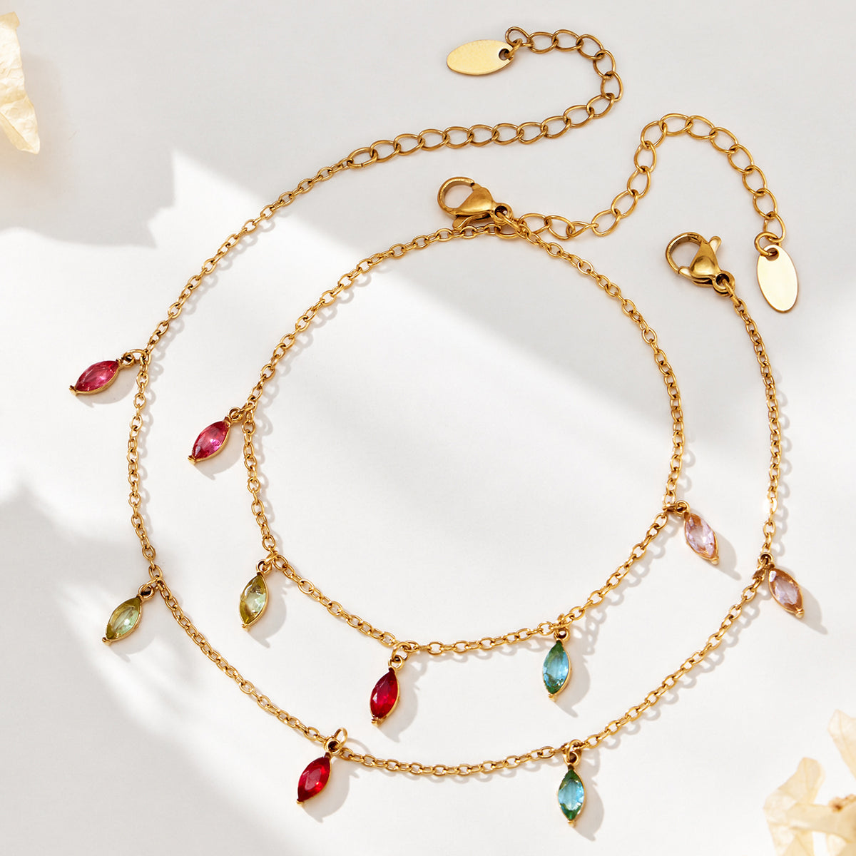 Two gold-tone fine-chain jewelry pieces with colorful teardrop charms and extension chains on a light surface.