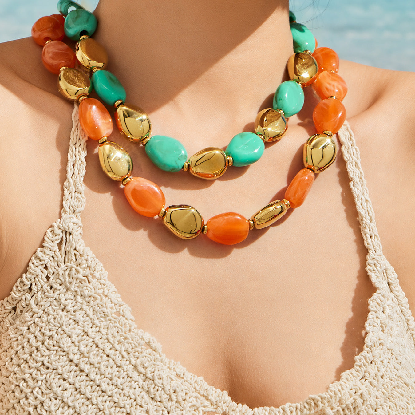 Model wearing layered orange-toned and green resin bead necklaces with polished gold-tone oval accents