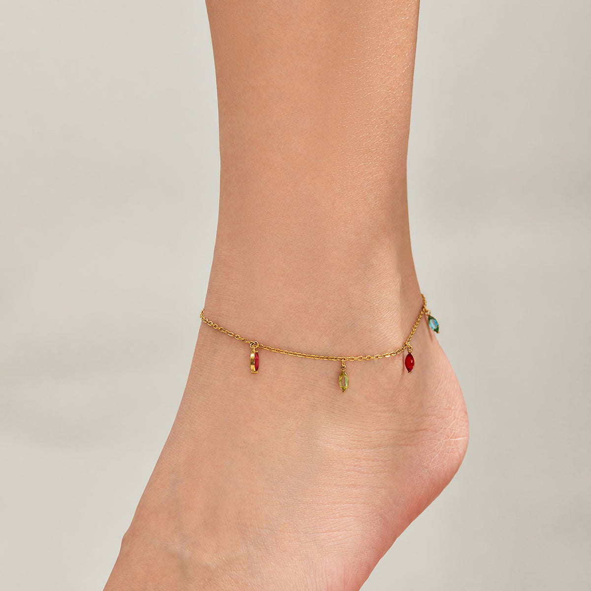 Side view of a delicate gold-tone anklet with small colorful teardrop charms around an ankle.