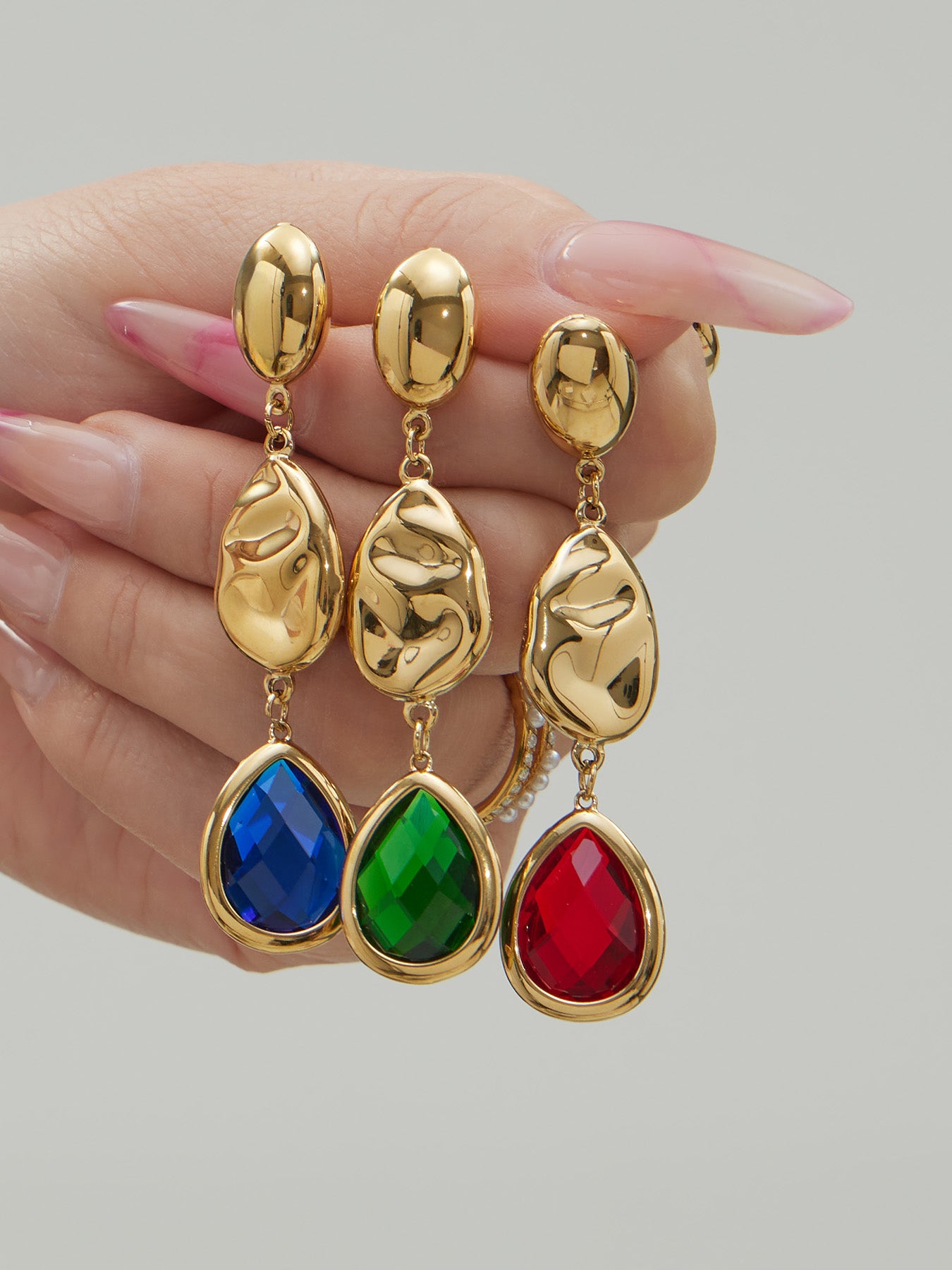 Hand displaying waterproof 316L stainless steel hammered gold earrings with emerald, sapphire, and ruby Czech crystals.