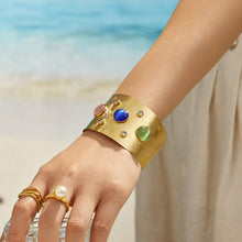 Wide gold cuff bracelet with colorful stones styled on a model hand by the sea